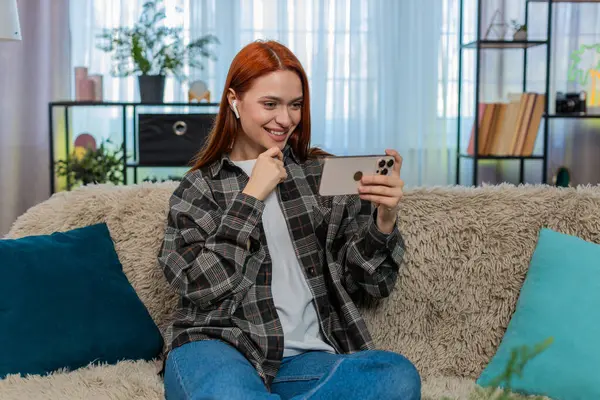Genç bir kadın akıllı telefondan video izleyip kulaklık takıyor ve güçlü duygularla gülüyor. Kafkasyalı kızıl saçlı kız kanepenin üzerinde eğleniyor, neşe ve neşeyle tepki veriyor.