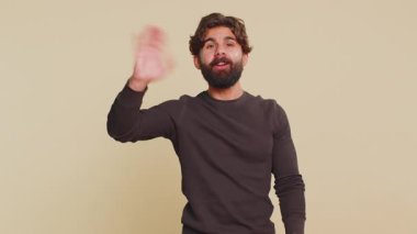 Merhaba, hoş geldiniz. Uzun kollu yakışıklı Hintli genç adam avucuyla selam veriyor, misafirperver bir gülümsemeyle selamlıyor. Yetişkin erkek İspanyol, bej renkli stüdyo arka planında kapalı..