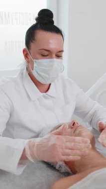 Güzellik uzmanı, genç yetişkin kadın yüzüne nemlendirici tıbbi kimyasallar soyarak krem fırçalama maskesi uyguluyor. Dişi cilt bakımı prosedürü. Yüz kozmetolojisi cilt yenilenmesi. Dikey