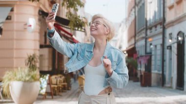 Neşeli genç bayan turist kameraya gülümsüyor, el sallıyor, el sallıyor merhaba, selam, selam, hoşçakal, misafirperver bir yüz ifadesiyle hoş geldiniz. Şehir sokağındaki sarışın kadın..