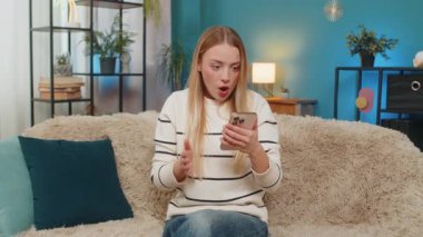 Beyaz bir kadın evdeki kanepede oturmuş online oyunu kutluyor ve zaferi kazanıyor. Heyecanlı kız akıllı telefonu tutuyor, sevinçle el kaldırıyor, gülümsüyor, mutluluğu, heyecanı ve başarıyı içeride hissediyor.