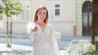 Gibi. Mutlu genç bir kadın kameraya onaylayıcı bir şekilde bakıyor ve olumlu bir geri bildirim imzalıyor. Şehir merkezindeki güneşli caddede bir kız. Şehir yaşam tarzları açık havada.