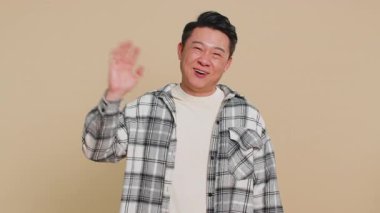 Kameraya gülümseyen mutlu Asyalı adam el sallayarak merhaba diyor ya da davetkar bir yüz ifadesiyle veda ediyor. Yakışıklı Çinli adam bej renkli stüdyo arka planında izole edilmiş.