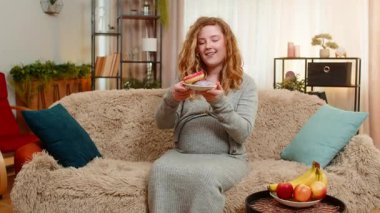 Hamile, genç, mutlu bir kadın elinde donutla kanepede oturuyor. Neşeli bir ifadeyle gülüyor, tatlı bir zevkin tadını çıkarıyor. Kız küçük zevkleri kucaklıyor, mutluluğu basit ve lezzetli anlarda buluyor..