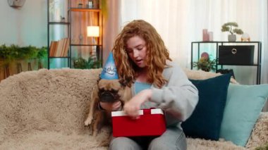 Beyaz saçlı genç bir kadın, doğum günü hediyesini sevinçle gülümseyerek ev kanepesinde açıyor. Pug dog kutuyu hevesle kokluyor, merakını belli ediyor ve sürpriz ve haz dolu anı paylaşıyor..