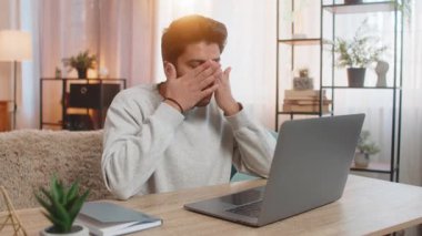 Genç Hintli adam dizüstü bilgisayarda çalışıyor, yorgun görünüyor, evdeki oturma odasındaki kanepede otururken gözlerini ovuşturuyor. İspanyol bir işkolik, görevine devam ediyor, bitkin görünüyor..