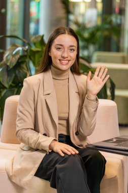 Ofis lobisinden uzaktan çekilmiş çevrimiçi konferans görüntülü konuşmasında gülümseyen serbest çalışan genç iş kadınının portresi. Koridorda dizüstü bilgisayarı olan resmi elbiseli bir kız..