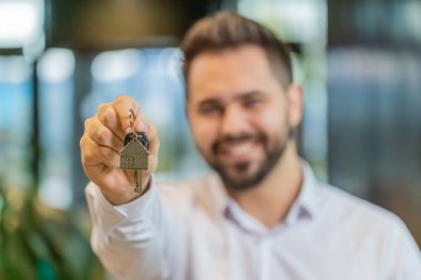 Mutlu genç emlakçı el kaldırıyor yeni ev dairesinin anahtarlarını gösteriyor, ev satın alıyor ya da kiralıyor modern ofis lobisinde ipotek kredisi veriyor. Yönetici anahtarı tutarak gösterir.