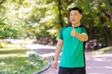 Gibi. Portre atletizm formundaki gülümseyen koşucu eğitimi maraton başarısından dolayı mutlu görünüyor. Parkta egzersiz kardiyolojisi. Japon genç sporcu bulanık ağaçlara karşı baş parmağını kaldırıyor