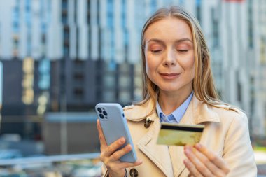 Kafkas 'lı yetişkin iş kadını kredi kartı ve akıllı telefon kullanarak internet üzerinden alışveriş siparişi veriyor. Kız şehir merkezindeki binaya karşı..