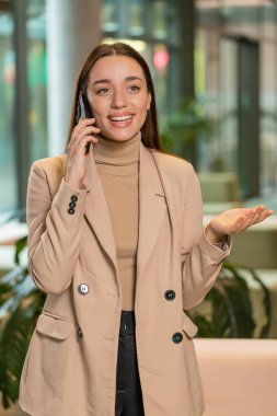 Resmi görevde gülümseyen genç bir iş kadını, modern ofis lobisinde cep telefonuyla konuşuyor. Ulak sohbet uygulamaları kullanarak akıllı telefonu tutan profesyonel yönetici kadın. İstihdam, meslek.