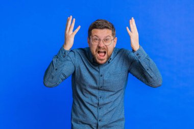 Kafasına dokunup kafa patlaması gösteren, endişeli ve şok olmuş, profesyonel tükenmiş, gözlüklü, orta yaşlı, heyecanlı bir beyaz adam. Genç yetişkin adam mavi arka planda izole edilmiş