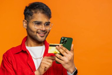 Kredi kartı ve akıllı telefon kullanan mutlu Hintli adam internet üzerinden alışveriş yaparken ve yemek siparişi verirken para harcıyor. Arap adam turuncu stüdyo arka planında izole edilmiş. Kopyalama alanı