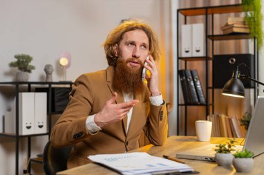Genç işadamı ofis masasında oturuyor cep telefonuyla müşteriye cevap veriyor, akıllı telefon kullanıyor. Sohbet. Kafkasyalı kızıl saçlı, uzaktan kumandalı bir işte çalışıyor..