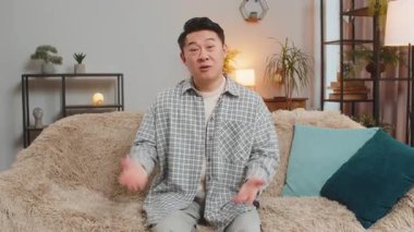 Mutlu orta yaşlı Çinli adam kameraya gülümsüyor ve el sallıyor merhaba, selam veriyor, evdeki oturma odasındaki kanepede otururken konuşuyor. Mutlu adam evde vlog kaydediyor.