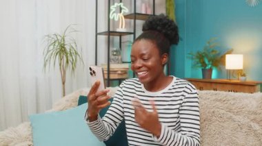 Afro-Amerikalı bir kadın akıllı telefondan birini selamlıyor. Evdeki kanepede oturmak, sıcak bir şekilde gülümsemek, el sallamak, mutluluğu ifade etmek, evdeki dijital bağlantıyla neşeli etkileşimi sürdürmek.