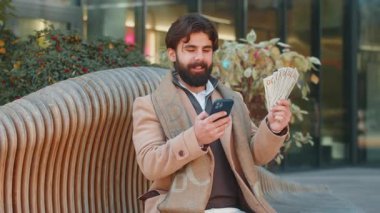 Akıllı telefon kullanan ve paralı banknotları sayan mutlu Hintli işadamı şehir merkezindeki bankta oturuyor. Başarılı iş kariyeri, piyango kazananı, büyük gelir, zenginlik