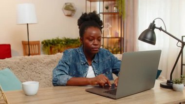 African American young woman sitting at table on home sofa flirts, shows playful call me hand gesture toward the camera with a sweet smile. Black girl looks confident expressive, enjoying the moment