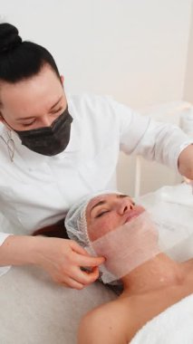 Kozmetoloji kliniğinde yüz bakımı gören orta yaşlı bir kadın. Güzellik uzmanı karboksiterapiye başlamak için nemlendirilmiş cilde gazlı bez uyguluyor. Hidrasyon, ışıltı, ton ve gençleştirme etkisini teşvik ediyor..