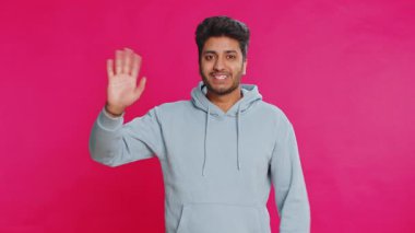 Kameraya dostça gülümseyen genç Hintli adam el sallayarak merhaba diyor ya da davetkar bir yüz ifadesiyle veda ediyor. Arap Hindu adam pembe stüdyo arka planında izole edilmiş.