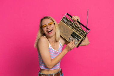 Otuz yaşında bir kadın, müzik dinlemek için retro teyp kullanıyor, disko dansı favori şarkısını dinliyor, eğleniyor, eğleniyor, klasik teknolojilerin hayranı. Pembe arka plandaki sarışın kız.