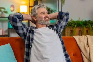 Gri saçlı, kollarını esneten, oturma odasındaki kanepede dinlenen yorgun, yaşlı bir adam. Düşünceli, orta yaşlı, kanepede uzanmış ellerini başının arkasına koyup eve bakan beyaz adam.