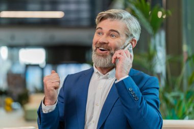 Takım elbiseli, akıllı telefon ve dizüstü bilgisayar kullanarak müşteriye cevap veren mutlu bir iş adamı. Sohbet. İnternette çalışan beyaz sakallı adam modern ofis lobisinde konuşuyor..