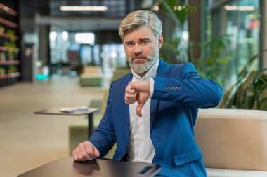 Hoşlanmamak. Takım elbiseli üzgün işadamı ofis lobisinde oturmuş işaret dilini gösteriyor hoşnutsuzluğunu, memnuniyetsizliğini ve kötü işini dile getiriyor. Otel odasında ciddi bir olgun adam var..