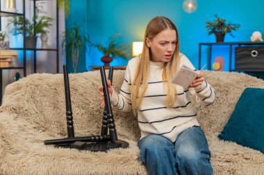 Beyaz kadın, evinde koltukta oturan yazılı talimatlarla küçük bir masa kurmaya çalışıyor. Kafası karışmış, endişeli, biraz hayal kırıklığına uğramış, kararsız, elinde kağıt bir diyagram, kararsız.