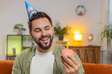 Mutlu adam elinde kek tutan neşeli bir doğum günü şapkası takıyor pastaya mum üflediği için sevinçle tebrik ediyor. Genç esmer adam doğum günü partisini sınıftaki kanepede kutluyor..