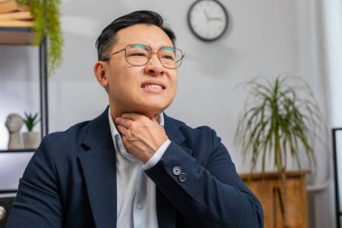 Asyalı, olgun iş adamı, boğazı ağrıdığı için hastalandığını, iş yerinde çalışırken boynuna dokunan ağrıyı yuttuğunu hisseder. Resmi takım elbiseli erkek Çinli nezle, grip ve virüs kapmış.