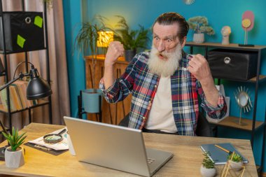 İç işlerinde kıdemli yaşlı bir adam dizüstü bilgisayar çığlığıyla zaferle el ele tutuşup başarıyı kutluyor piyangodan para kazanıyor. Heyecanlı yaşlı büyükbaba internetten iyi haberler alıyor..