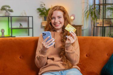 Para transfer ederken kredi kartı ve akıllı telefon kullanan mutlu genç bir kadın. Evin içinde alışveriş yaparken nakit para almadan yemek siparişi veriyor. Kanepede oturan kızıl saçlı kız..