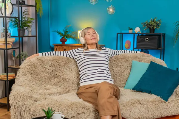 Beyaz, olgun bir kadın, evdeki kanepede rahatça oturuyor. Mutlu büyükanne gözlerini kapatır ve kablosuz kulaklıklarla sakin müzik dinlemeyi sever. Rahat ve huzurlu görünürken rahat bir odada dinlenir.