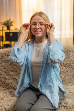 Genç bir kadın evde şık gözlük çerçeveleri takarak günlük işler için kendine güven ve netliği arttırıyor. Kanepede oturan beyaz kız, sıcak gün ışığında göz desteğiyle birleştiriyor..