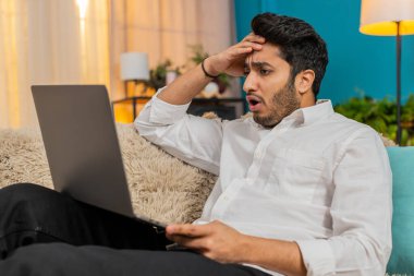 Hintli adam evde hayal kırıklığına uğramış hissediyor, online oyun kaybına tepki veriyor, hayal kırıklığı ve gerilim gösteriyor. Arap adam kanepede oturuyor, başarısız oyun denemesinden sonra stresli, dizüstü bilgisayara odaklanmış.
