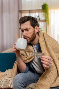Evdeki genç adam sıcak bir battaniyeye sarılı olarak kanepede oturuyor, kendini daha iyi hissetmek için elinde bir fincan çay tutuyor. Kafkasyalı adam iyi görünmüyor, masada ilaç var, muhtemelen hastalıktan ya da soğuktan iyileşiyor.