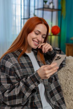 Genç, mutlu bir kadın akıllı telefondan haber aktarıyor. Hoş bir gülümsemeyle ara sıra güncellemelere tepki veriyor. Kanepedeki beyaz saçlı kız sosyal medya içeriğinin tadını çıkarıyor, rahat, neşeli bir an.