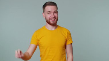 Sarı tişörtlü yakışıklı adam zafer işareti gösteriyor, başarı ve kazanmayı umuyor, barış jesti yapıyor, iyimser bir ifadeyle gülümsüyor. Gri duvar arkasında yalnız genç bir adam.