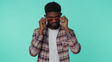 20 yaşında, kırmızı güneş gözlüğü takmış, gülümseyen, göz kırpan, göz kırpan iyimser bakışlarıyla kameraya bakan, göz kırpan ve flört eden neşeli Afro-Amerikan bir adam. Mavi arka planda genç bir yetişkin.