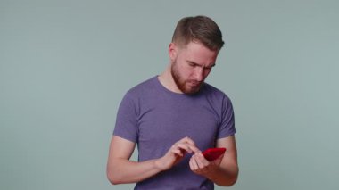 Gayri resmi tişörtlü yakışıklı yetişkin adam cep telefonu kullanıyor web mesajlarına yeni mesajlar yazıyor, internette geziniyor, sosyal ağlara bağımlı oluyor. Stüdyoda çekilen genç adam gri duvar arkasında tek başına.