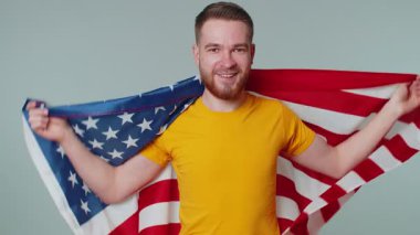 Tişörtlü, Amerikan bayrağına sarılı sakallı bir gencin portresi. Kutlama, insan hakları, özgürlükler. Bağımsızlık Günü. Stüdyoda çekilen yetişkin adam gri arka planda tek başına.