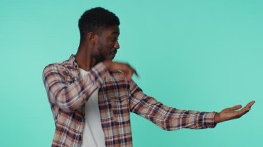 Kareli tişörtlü yakışıklı Afro-Amerikan adam. Başparmaklarını kaldırıyor, boş yeri işaret ediyor. Ticari metinler için reklam alanı, ürün tanıtımı için kopya alanı. Mavi arka planda çekilen kapalı stüdyo.