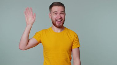 Tişörtlü yakışıklı adam kameraya gülümsüyor ve el sallayarak merhaba ya da güle güle diyor, misafirperver bir yüz ifadesiyle karşılıyor. İçerideki mutlu genç adam gri duvar arkasında izole edilmiş.