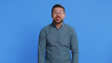 Beyaz, orta yaşlı, gözlüklü, dil gösteren, kameraya surat yapan, şakalaşan, aptal suratlı, şakacı, zorba, tacizci bir adam. Genç yetişkin adam mavi arka planda kapalı alanda izole edilmiş.