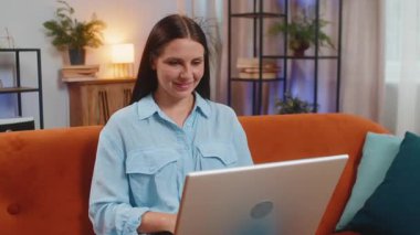 Oturma odasındaki işi bittikten sonra kanepeye oturmuş laptopu kapatan beyaz bir kadın. Mutlu genç kız serbest çalışıyor. Ev kanepesinde uzaktan kumandayla çalışıyor. Defter bilgisayarında e-öğrenme, internette gezinme