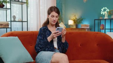 İnternetten alışveriş yapan mutlu genç bir kadın. Gülümseyen beyaz kız elinde bankacılık kredi kartı tutuyor evdeki koltukta mobil uygulama doğrulama sürecinde yer alıyor..