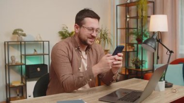 Masada oturan, cep telefonunu kullanan ve gülümseyen genç, beyaz bir adam. Guy Freelancer akıllı telefon sosyal medya uygulamaları üzerinden online film izlerken mesajlarını paylaşıyor.