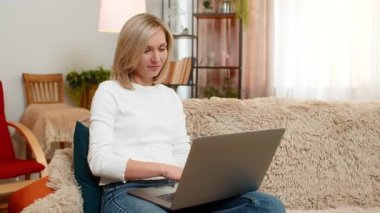 Orta yaşlı bir kadın, dizüstü bilgisayardaki görevi başarıyla bitirdikten sonra sevinçle evinde oturuyor. Yetişkin kadın iş başarısını, gururlu ve olumlu duygusal ifadeyle gülümseyerek kutluyor..