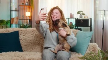 Genç beyaz blogcu kadın video görüşmesinde el sallıyor. Kanepede oturuyor, gülümsüyor ve neşeli görünüyor. Kız pug dog 'u tutarken teması selamlıyor, yanında evcil hayvanla sevimli bir anı paylaşıyor..